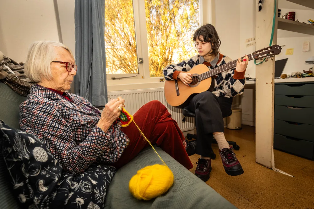 Een huurder speelt gitaar in zijn kamer terwijl de hospita breit